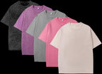Five plain t-shirts in black, purple, gray, pink, and beige arranged in a row on a white background.